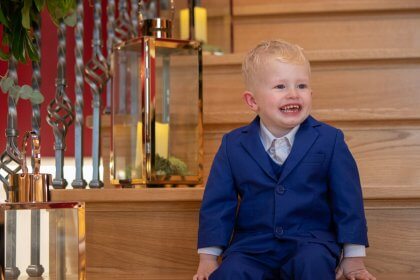 Smiling boy in blue suit sitting on stairs decorated with garland of flowers and candles in glass lanterns