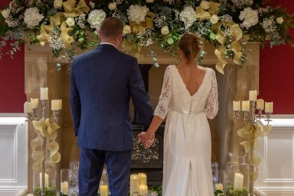 Close up of couple in wedding attire standing in front of decorated fireplace, viewed from behind